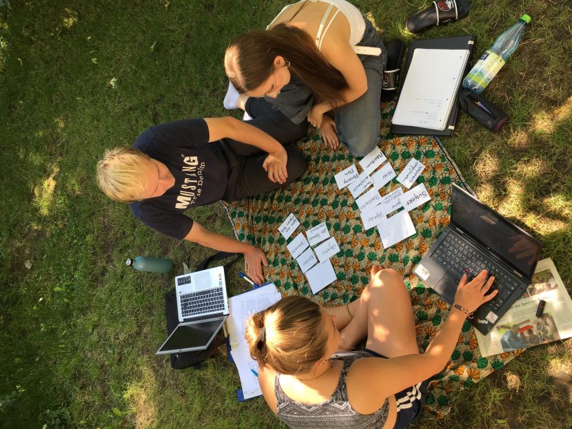 Nils, Lara und Lexi sitzen auf einer Decke im Gras mit ihren Laptops und haben Karteikarten vor sich ausgebreitet.