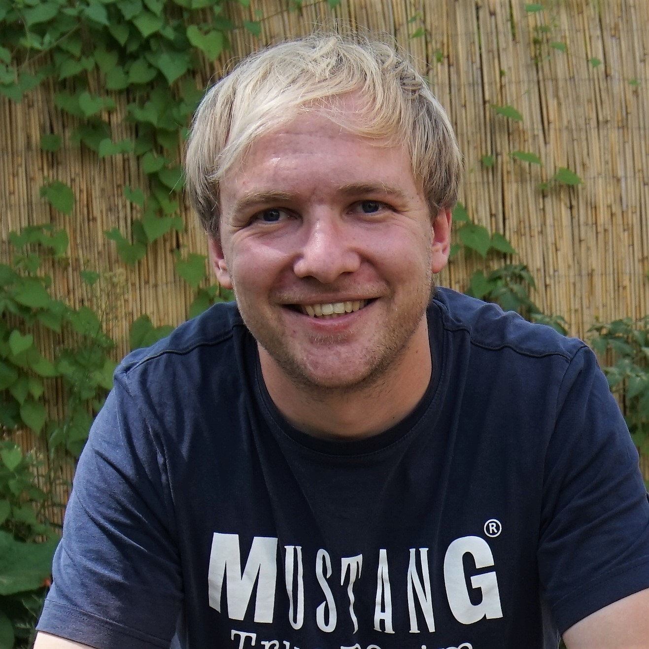 Ein junger Mann mit blonden Haaren sitzt vor einem Bambuszaun und blickt lächelnd in die Kamera